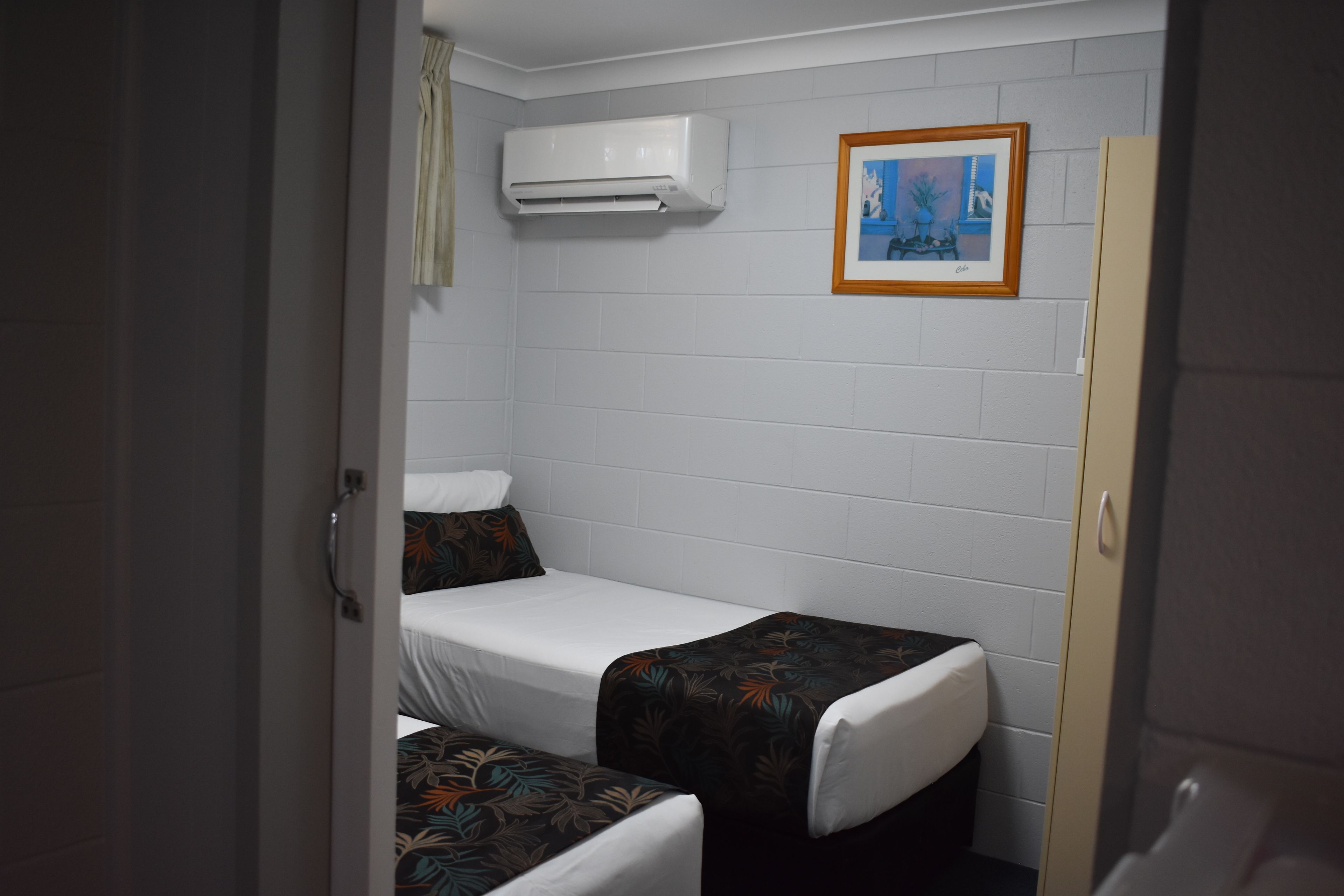 A small, compact room with two single beds, a white air conditioning unit, and a framed picture on the wall.