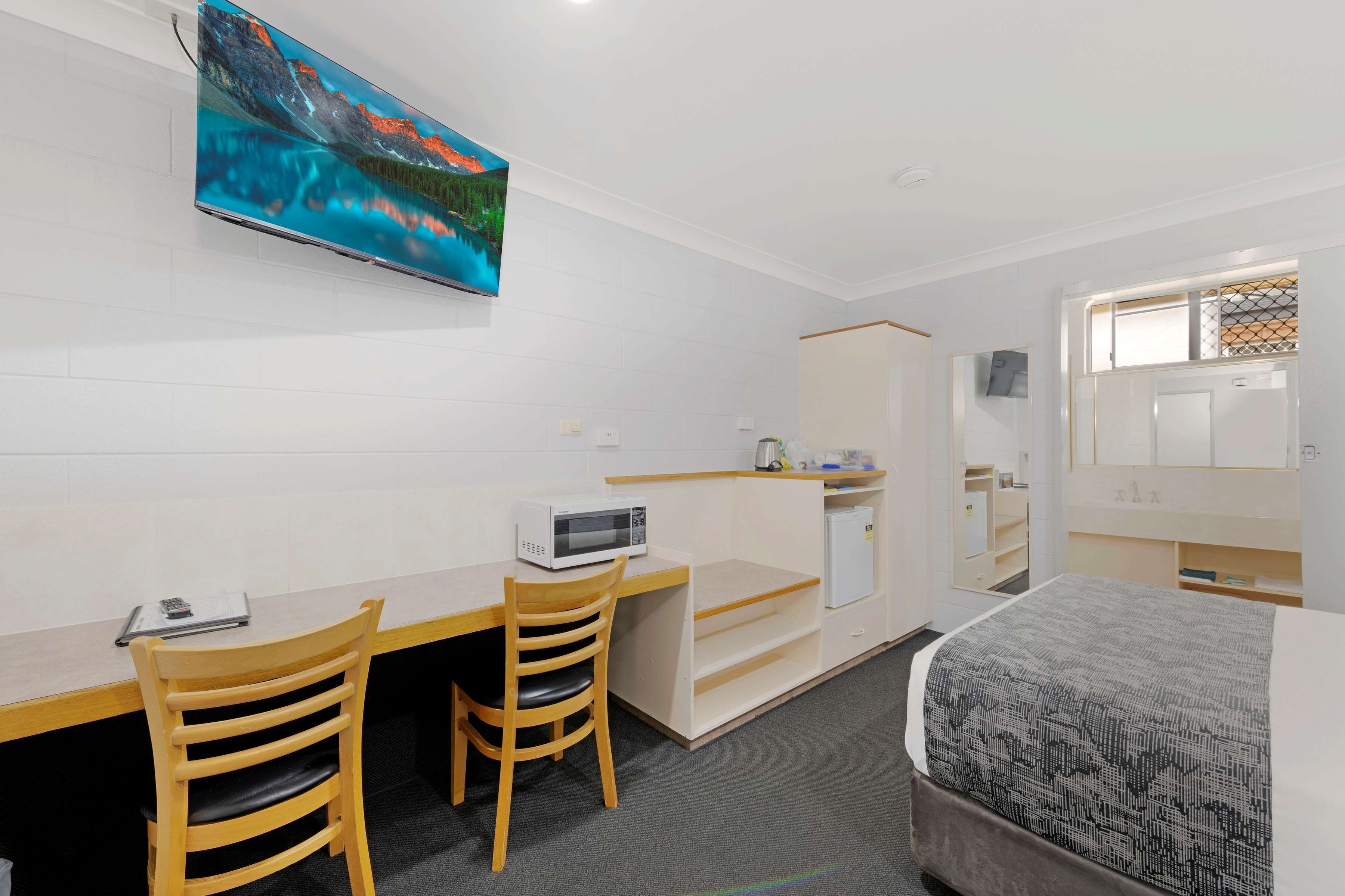 A hotel room with a bed, a desk with a chair, a microwave, a TV mounted on the wall, and a small kitchenette.