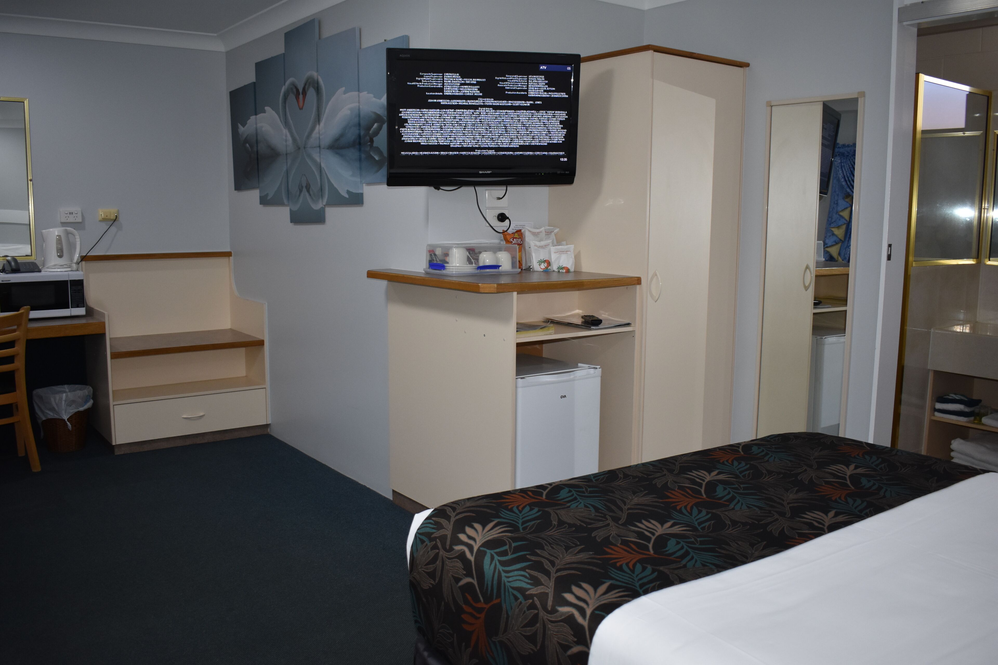 A hotel room with a bed, a desk with a computer, a television mounted on the wall, and a wardrobe.