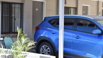 Um carro azul estacionado numa área coberta de estacionamento, com portas numeradas, uma planta em vaso e uma cadeira.