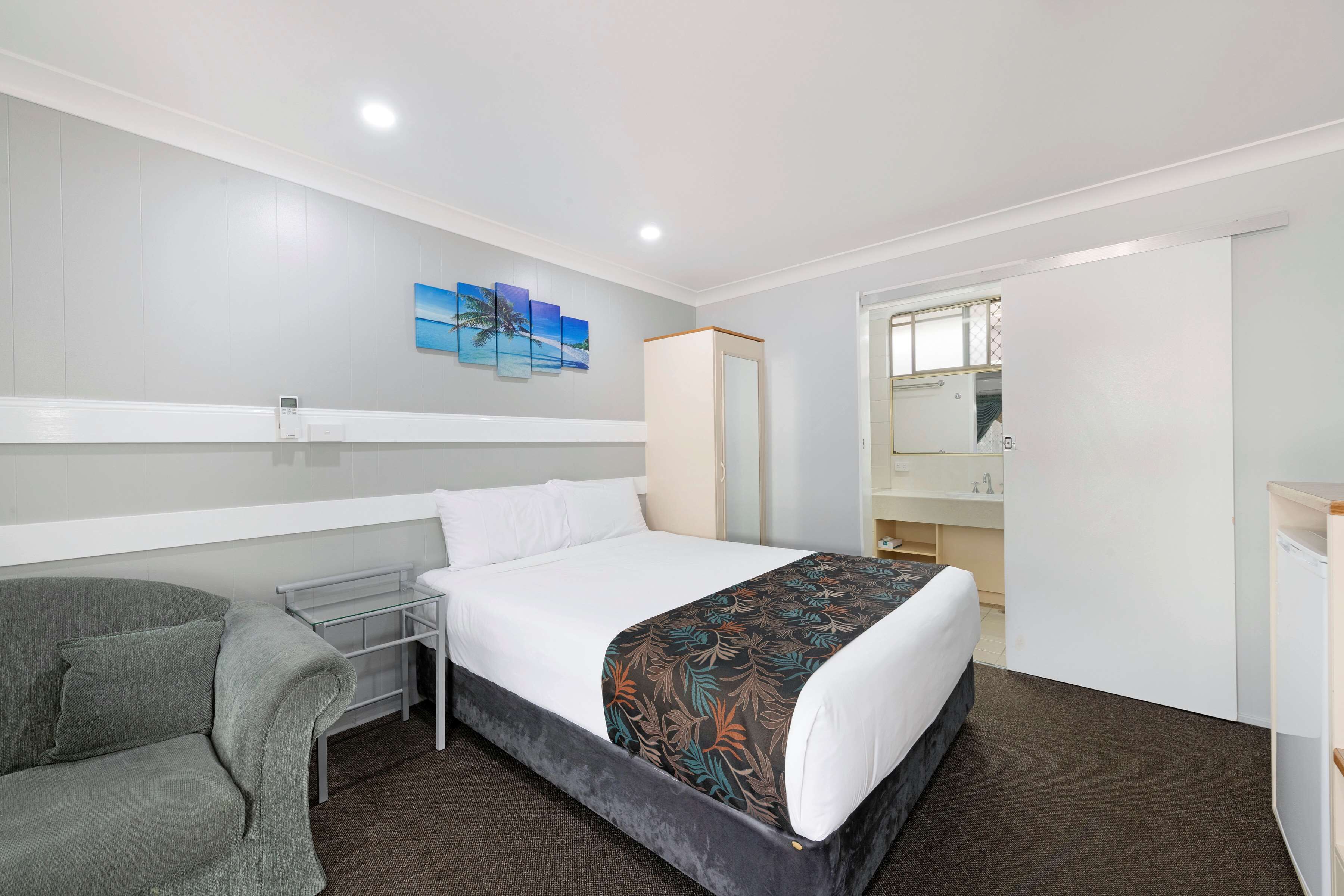 A hotel room with a bed, a gray sofa, a small table, a mirror, and a bathroom visible through an open door.