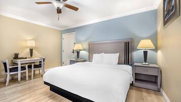 A hotel room with a bed, desk, chairs, lamps, a ceiling fan, and a door.