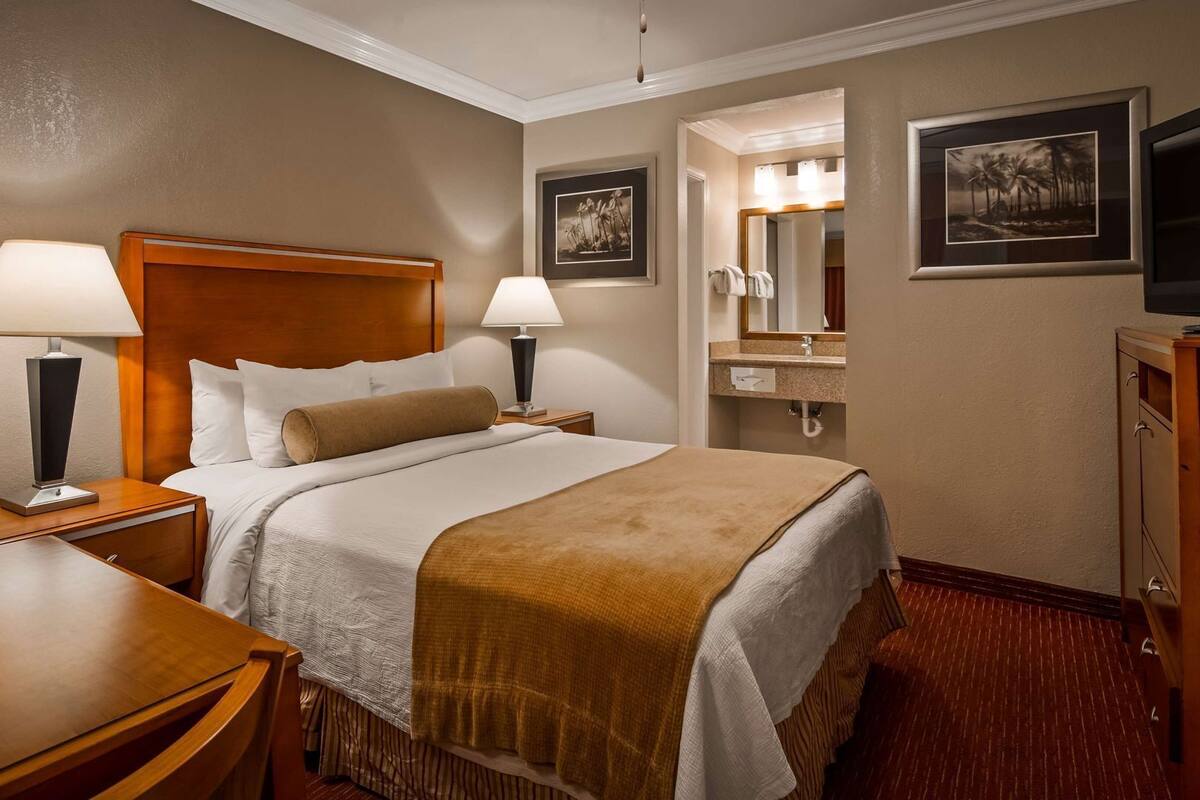 A hotel room with a bed, two bedside lamps, a desk, a chair, a television, and a bathroom with a sink and mirror.