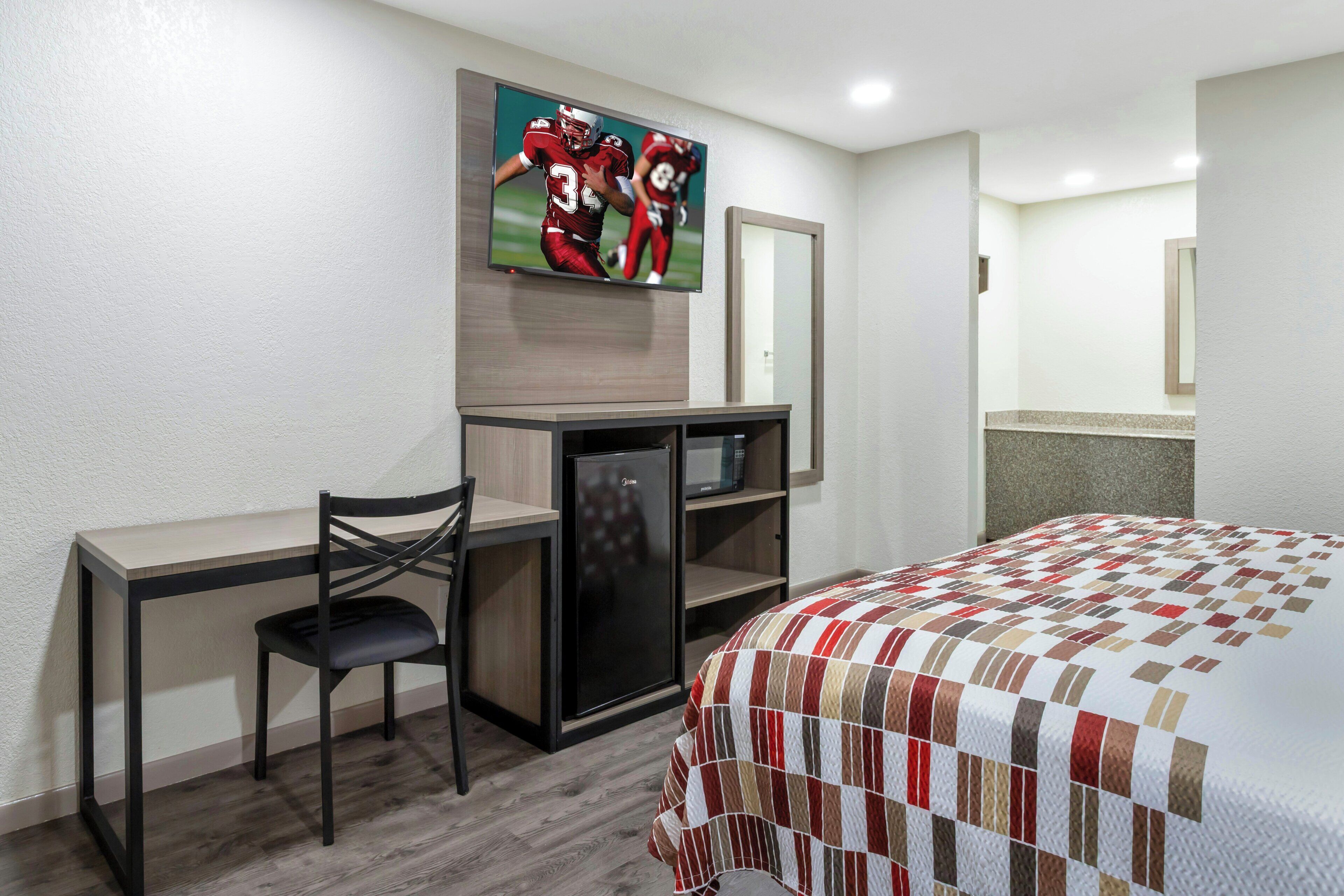 Habitación de hotel con cama, escritorio con silla, televisión montada en la pared y un refrigerador pequeño.