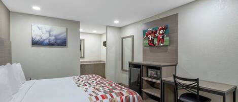 A hotel room with a bed, a desk, a chair, a refrigerator, and artwork on the walls.