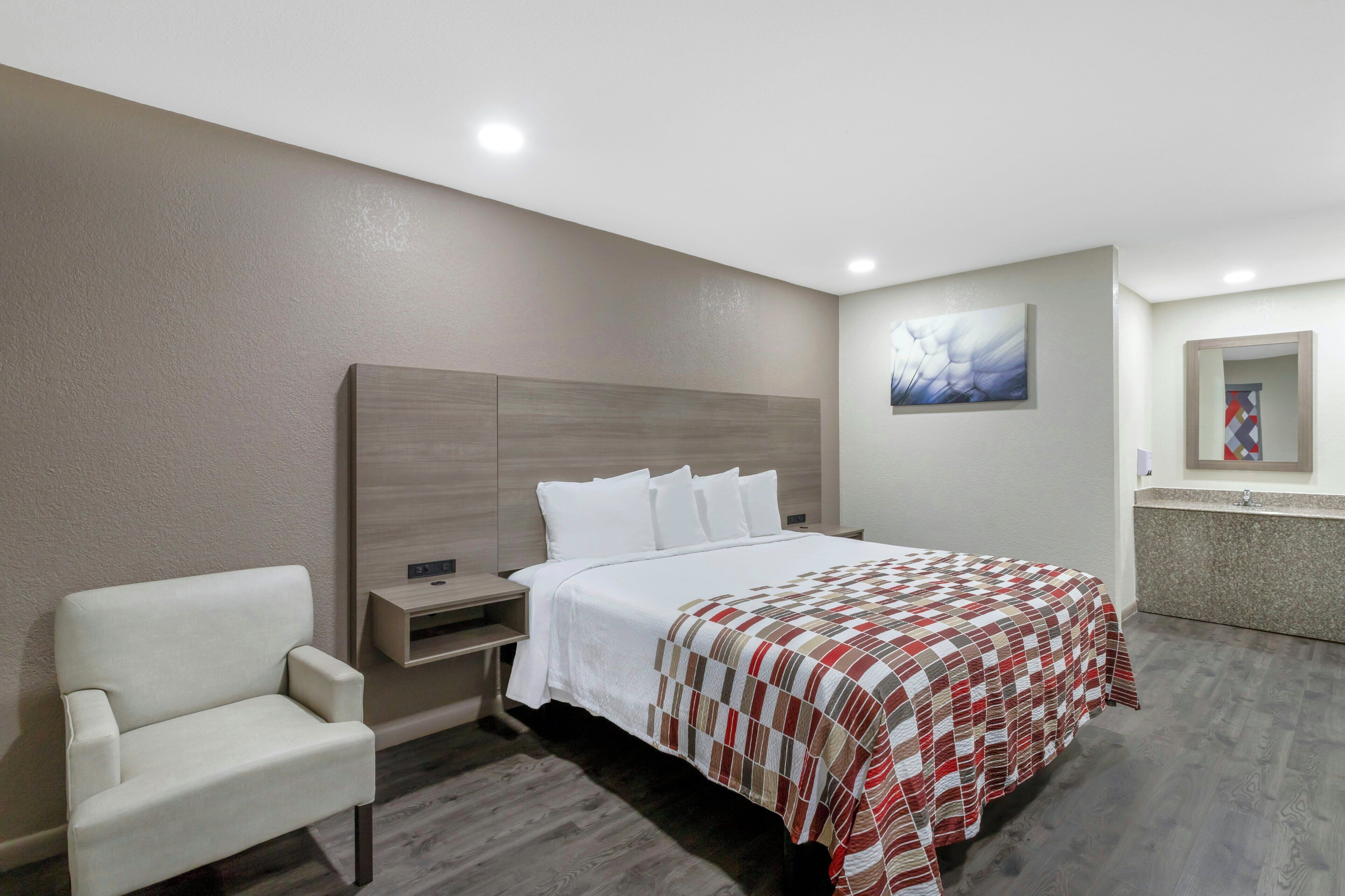 A hotel room with a bed, a chair, a nightstand, a painting, and a bathroom visible through an open door.