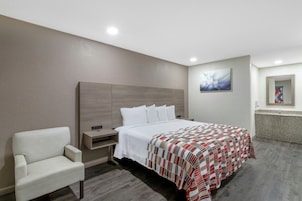 Habitación de hotel con cama, una silla, mesita de noche, un cuadro y baño visible a través de una puerta abierta.