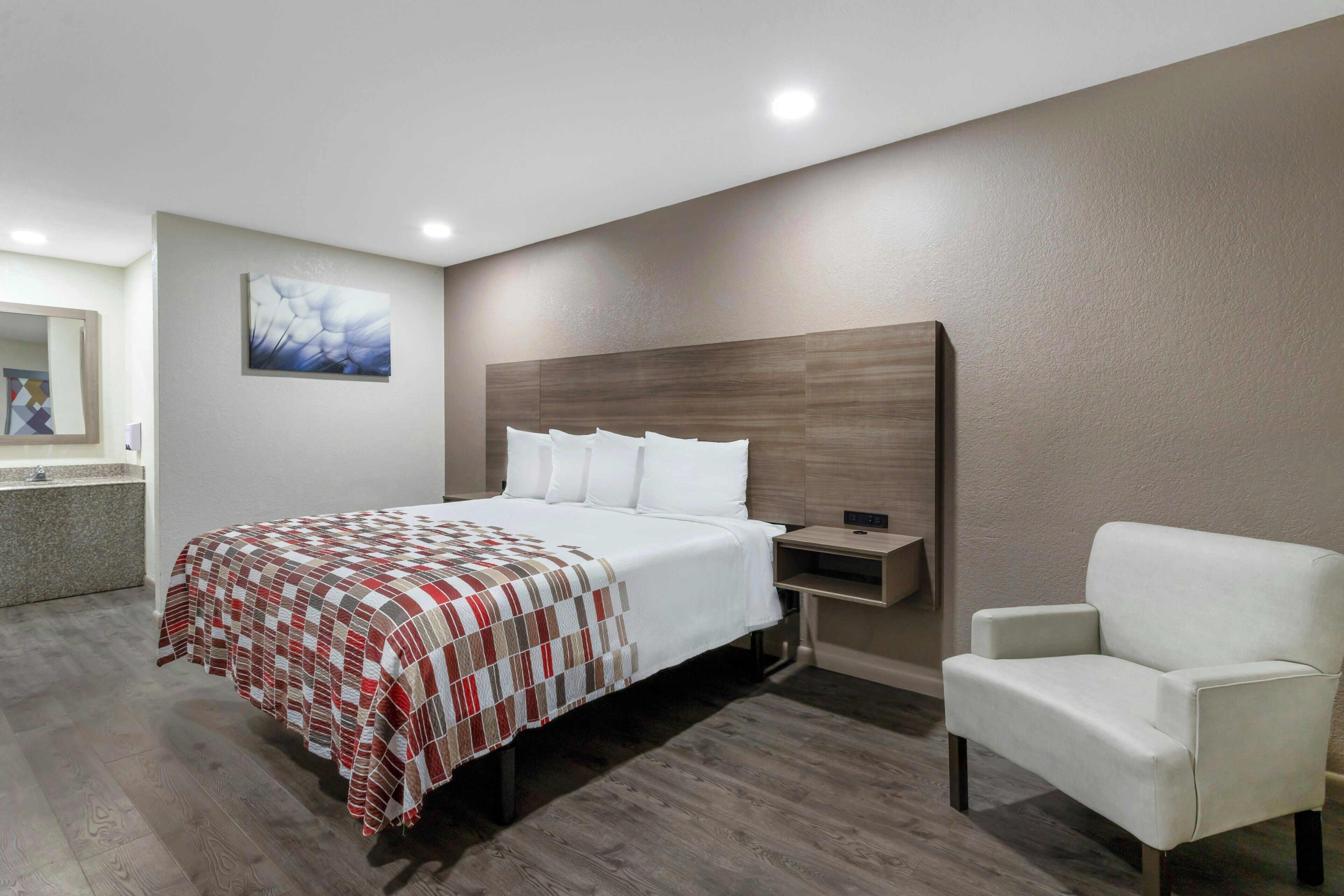 A modern hotel room with a large bed, a white armchair, a bedside table, and a wall-mounted artwork.