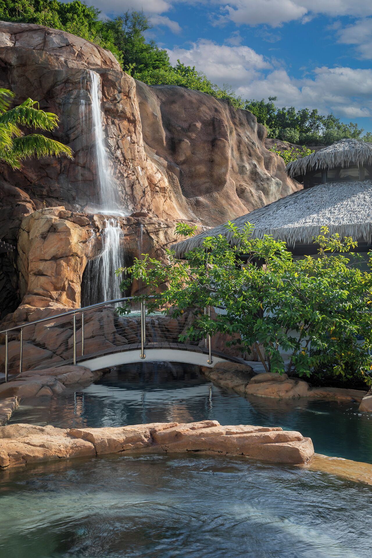 A waterfall cascading down a rocky cliff, a bridge over a calm pool, and lush greenery.