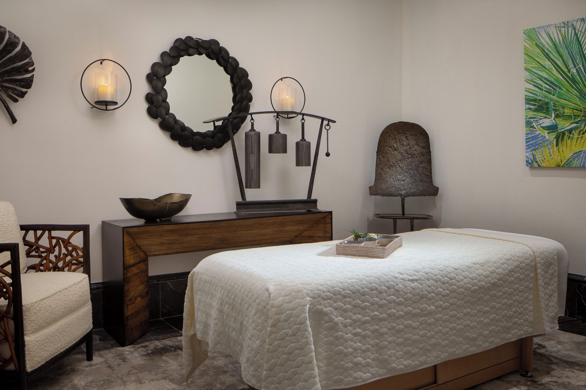 A bedroom with a bed, a wooden dresser, a chair, a desk, wall sconces, a mirror, and a painting.