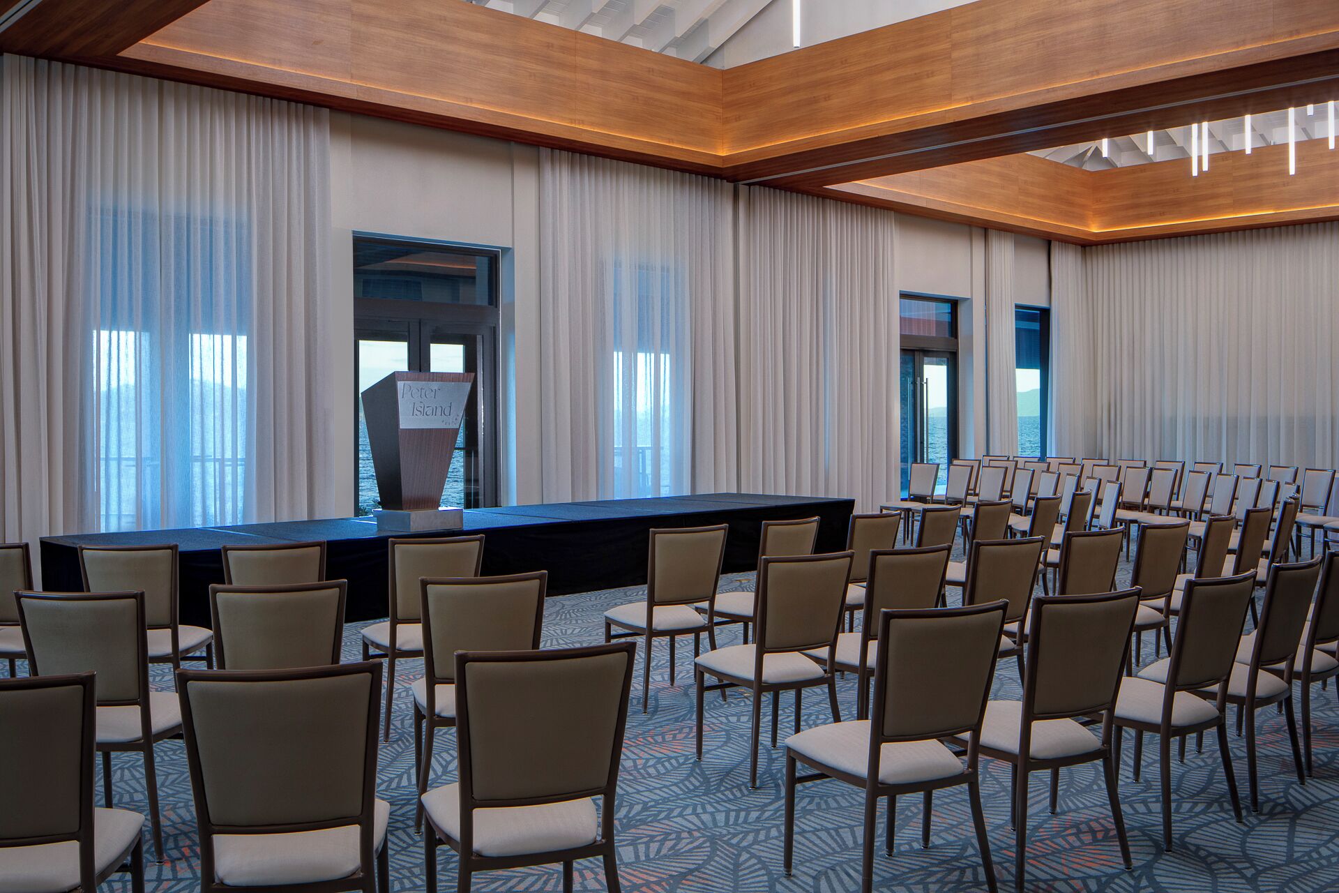A conference room with a long table, rows of chairs, and a view of the sea through large windows.