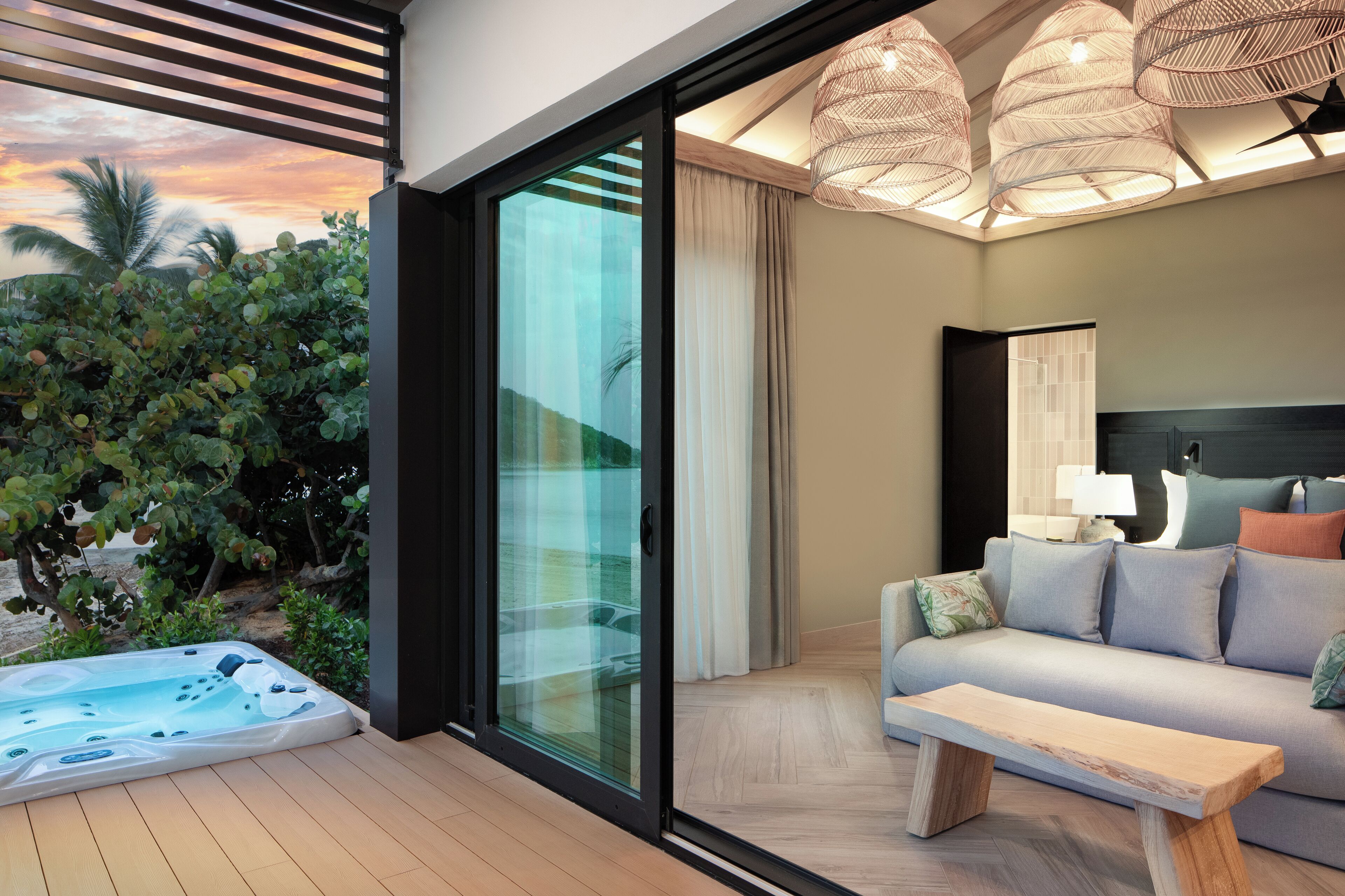 Une chambre moderne dotée d’une porte vitrée donnant sur un jardin, d’un canapé, d’une table de chevet et d’une grande fenêtre offrant une vue sur des palmiers.