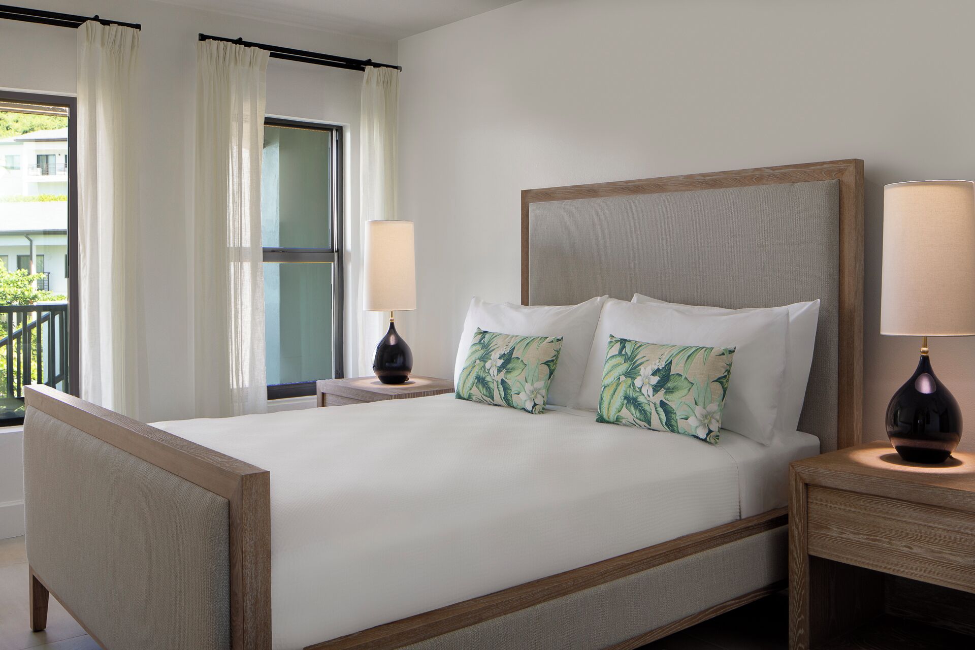 A hotel room with a large bed, two nightstands, a window with curtains, and a view of buildings outside.