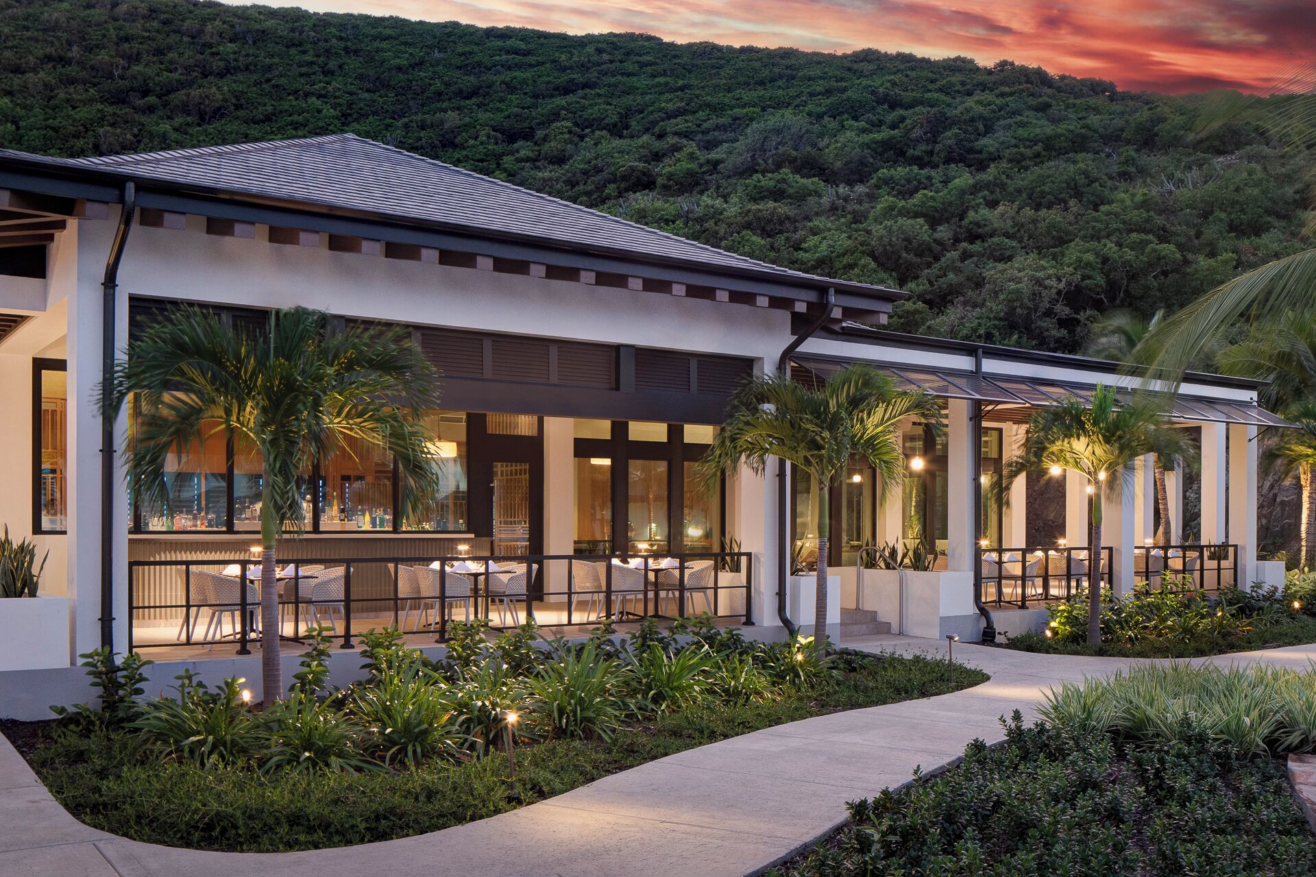 A modern building with a covered outdoor seating area, surrounded by lush greenery and palm trees.
