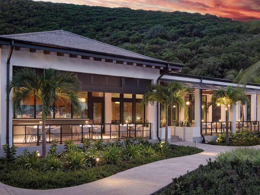 A modern building with a covered outdoor seating area, surrounded by lush greenery and palm trees.