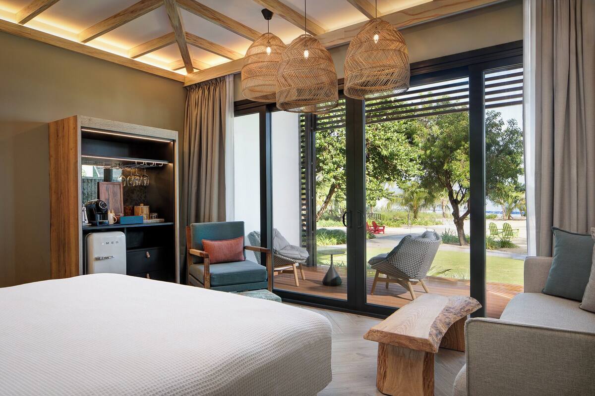 Une chambre d’hôtel moderne dotée d’un grand lit, d’un coin salon avec un banc et des fauteuils, d’un minibar et offrant une vue sur l’extérieur.