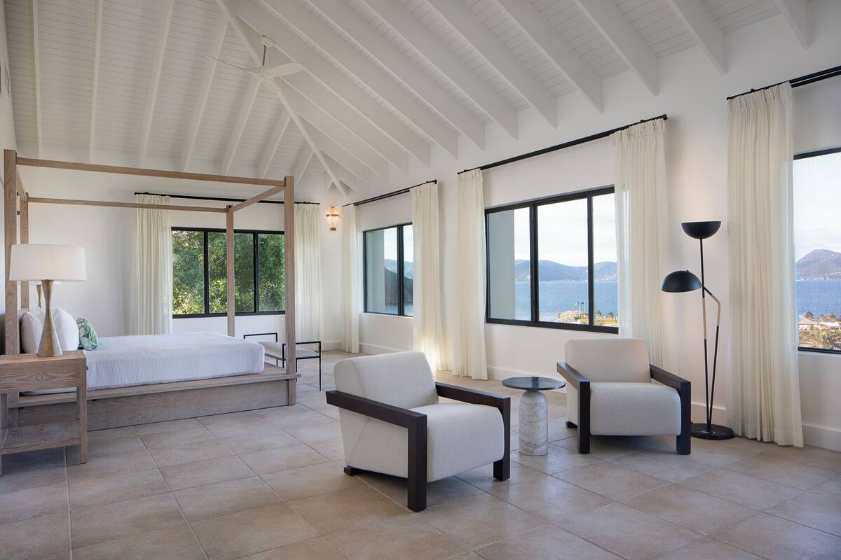 Une chambre spacieuse avec un lit à baldaquin, deux fauteuils, une petite table et de grandes fenêtres offrant une vue panoramique.