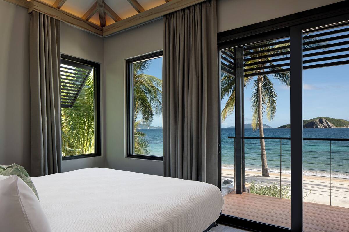 Une chambre avec un grand lit, deux fenêtres avec des rideaux et une vue sur l’océan et les palmiers.