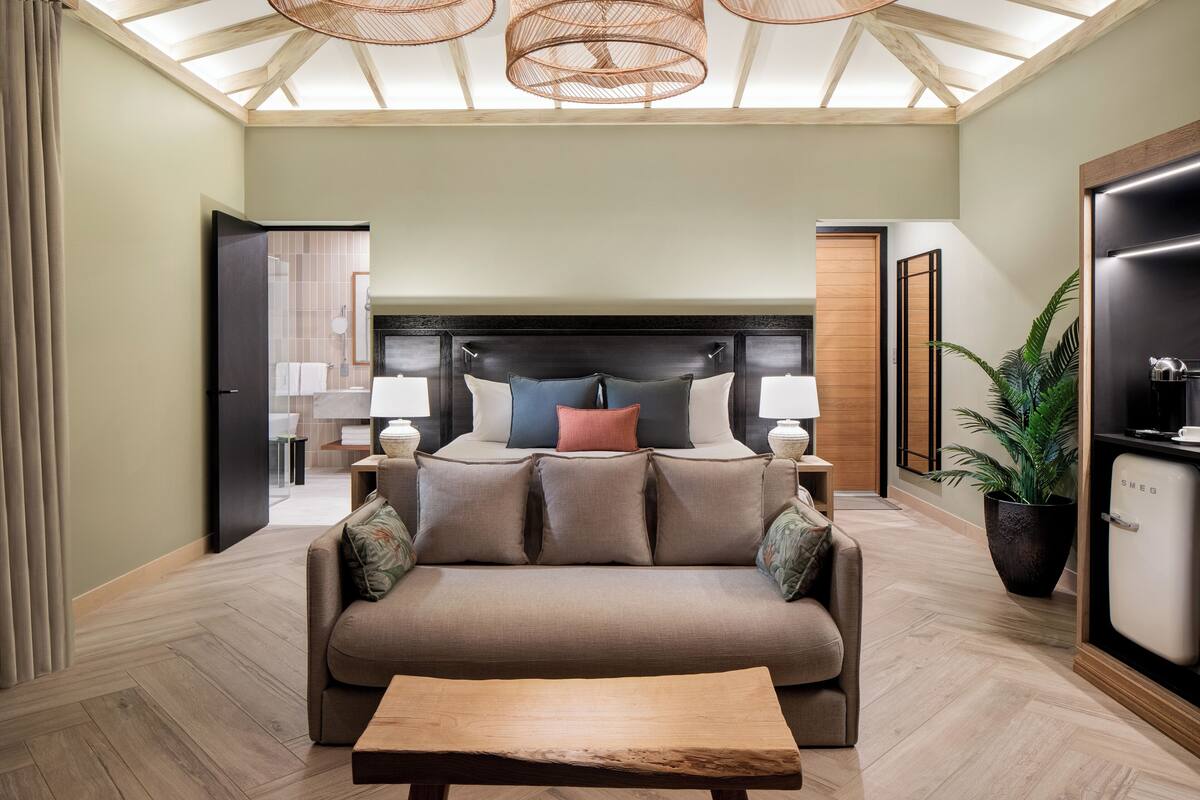 Une chambre d’hôtel moderne dotée d’un grand lit, d’un canapé, d’une table basse et d’un plafond avec des poutres en bois et des luminaires tressés.