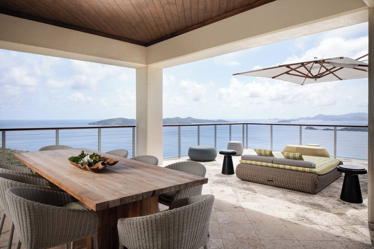 Une terrasse avec une table en bois, des chaises en osier, un canapé avec des coussins et un grand parasol.
