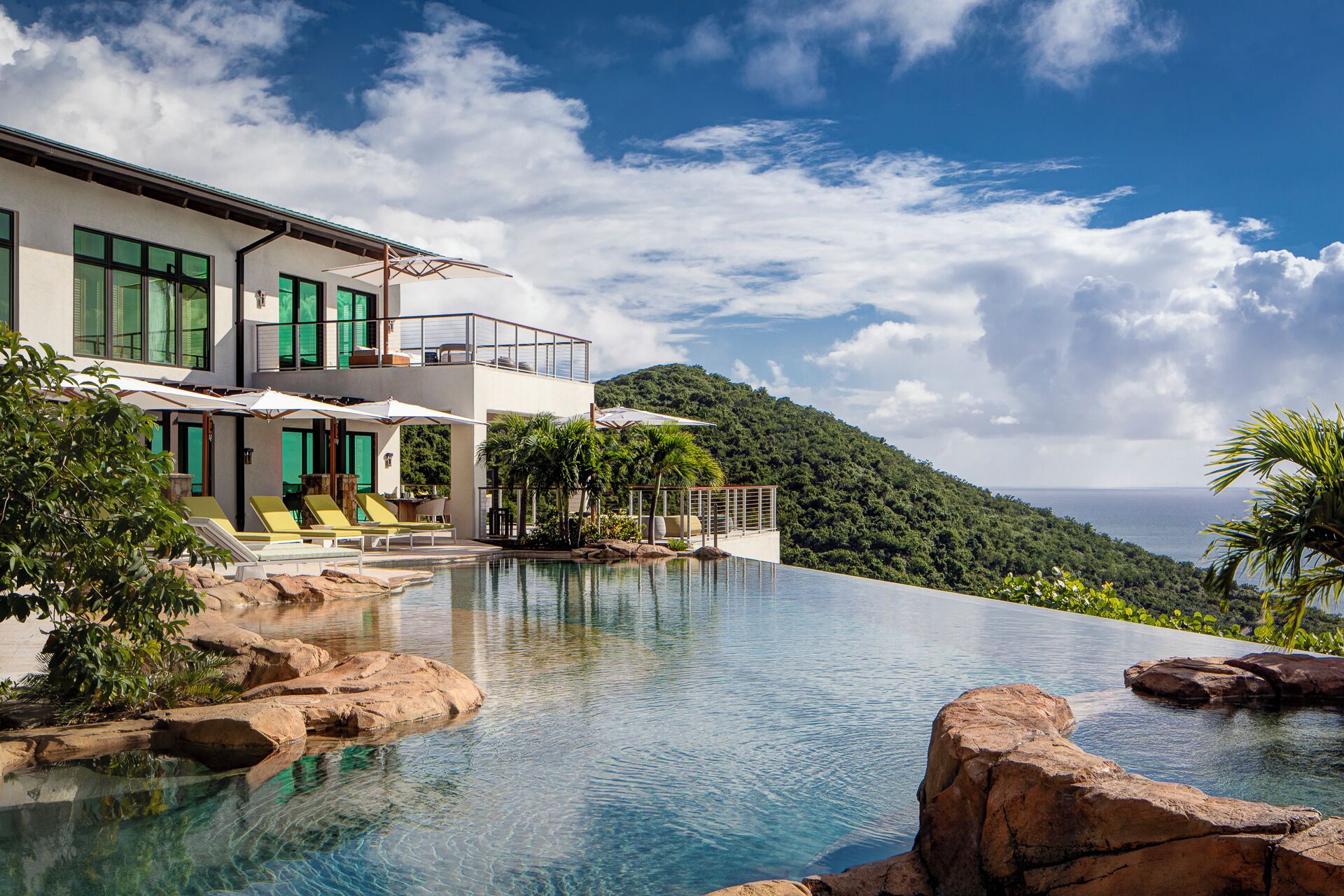 A modern villa with a private infinity pool, outdoor seating, and a scenic hillside view.