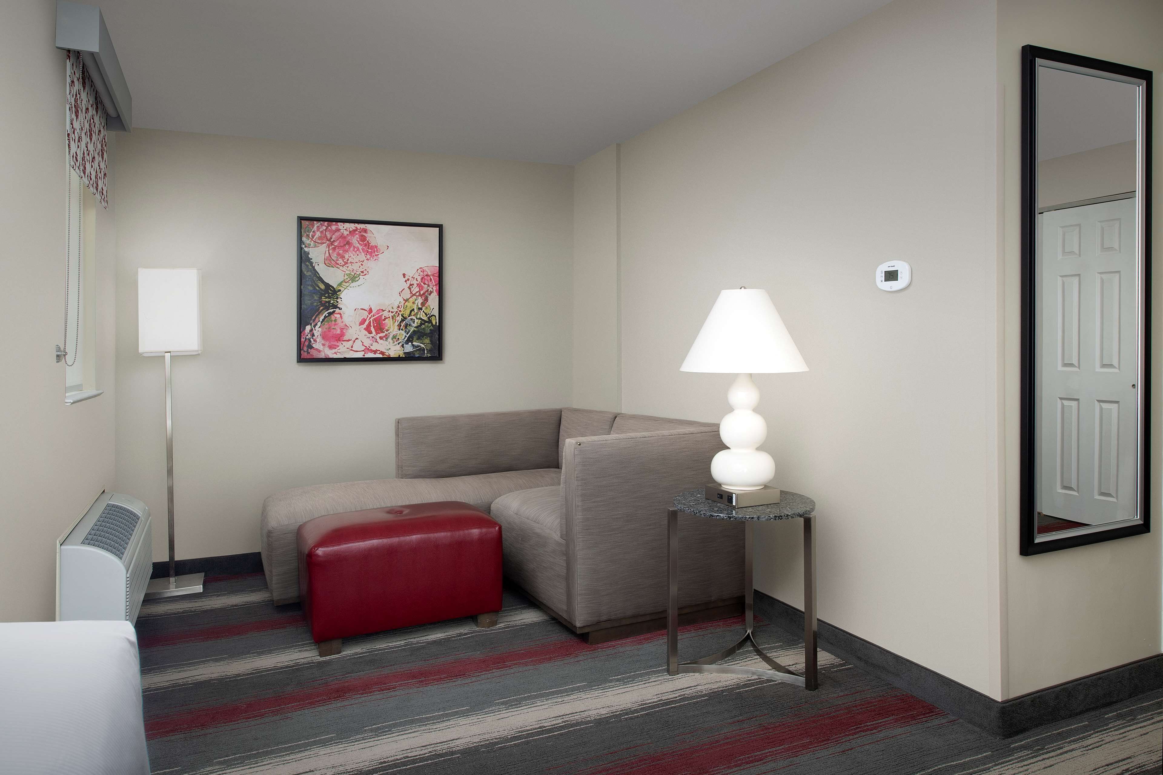 Habitación de hotel con sofá, un reposapiés rojo, lámpara de pie, una obra de arte floral enmarco y un espejo en la pared.