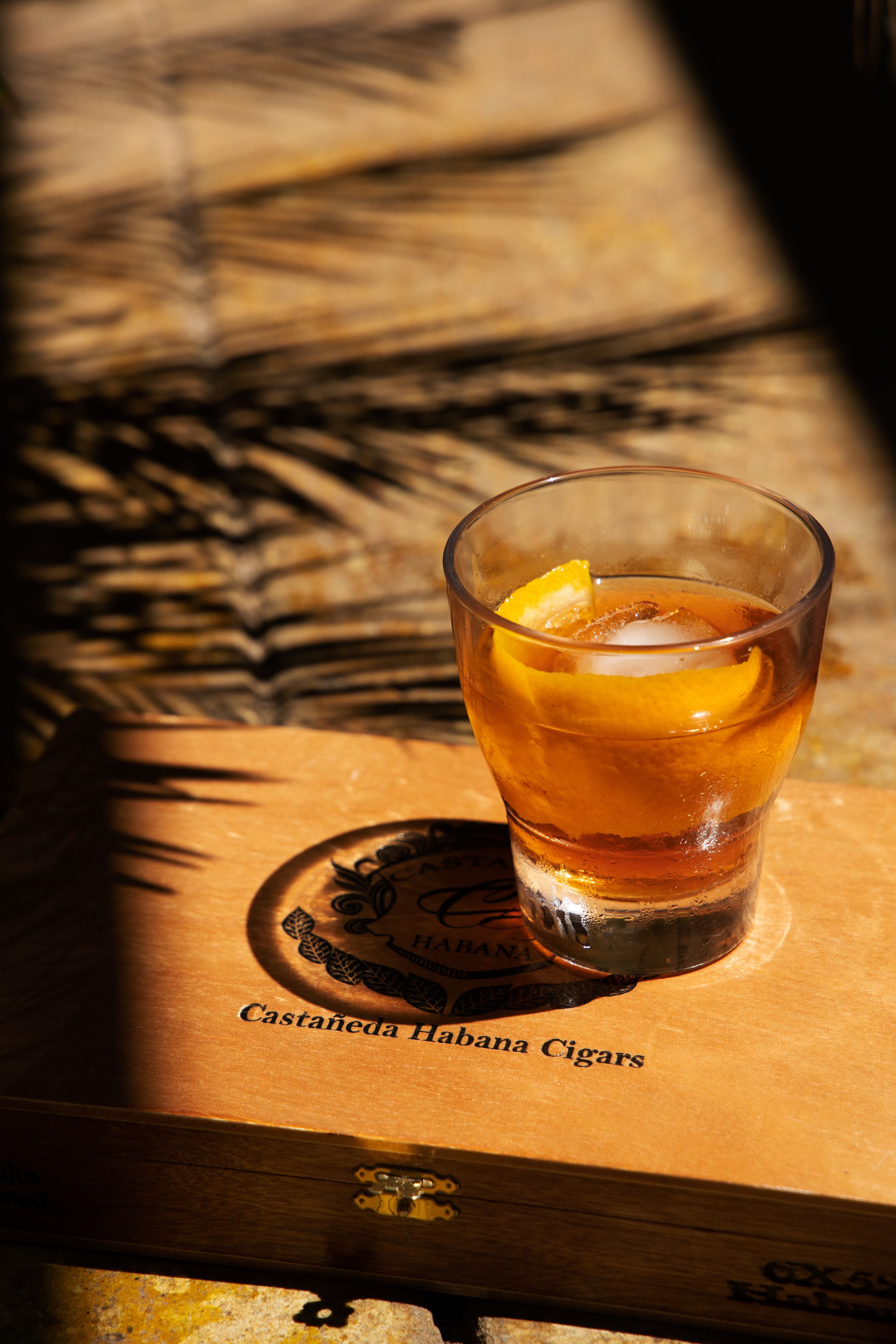 A glass of amber liquid with an orange slice, placed on a wooden surface with a logo and text reading "Castañeda Habana Cigars".
