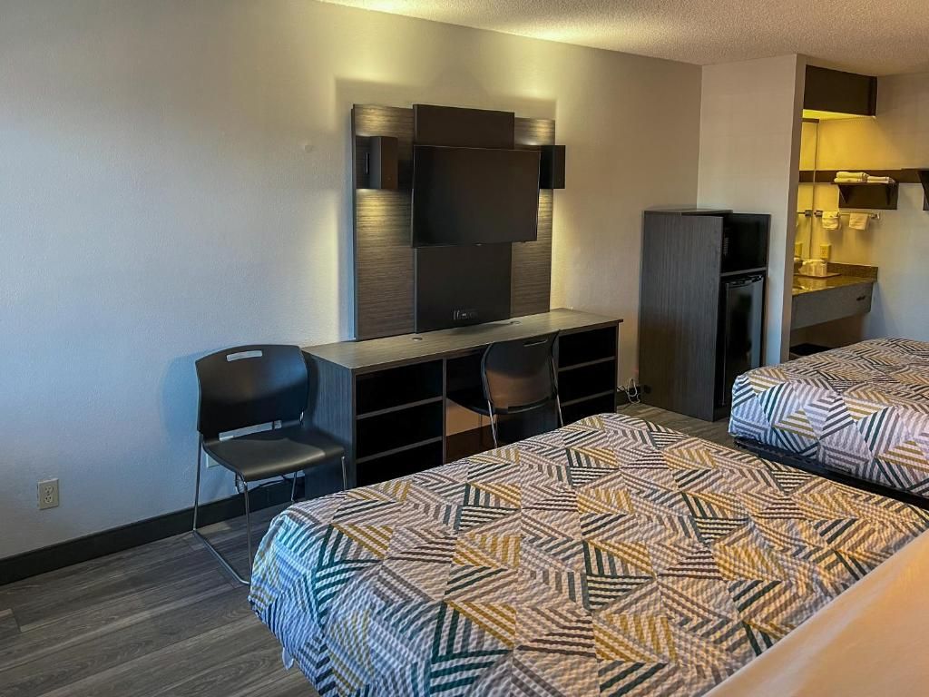 Quarto de hotel com duas camas, uma escrivaninha com cadeiras, televisão e guarda-roupa.