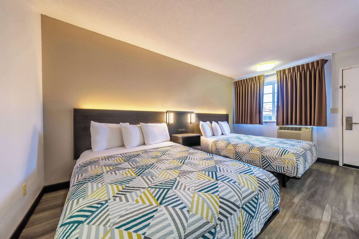 Une chambre d’hôtel avec deux lits, chacun recouvert d’une couvre-lit à motifs, une table de chevet, une lampe fixée au mur et une fenêtre avec des rideaux.