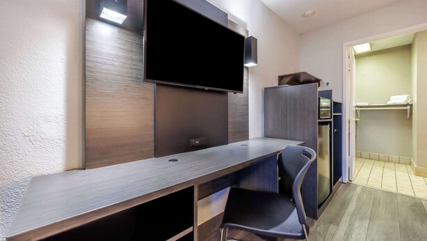A modern hotel room with a flat-screen TV mounted on the wall, a wooden desk with a chair, a mini-fridge, and a walk-in closet. - Motel 6 Portland, OR - Southeast (Portland)