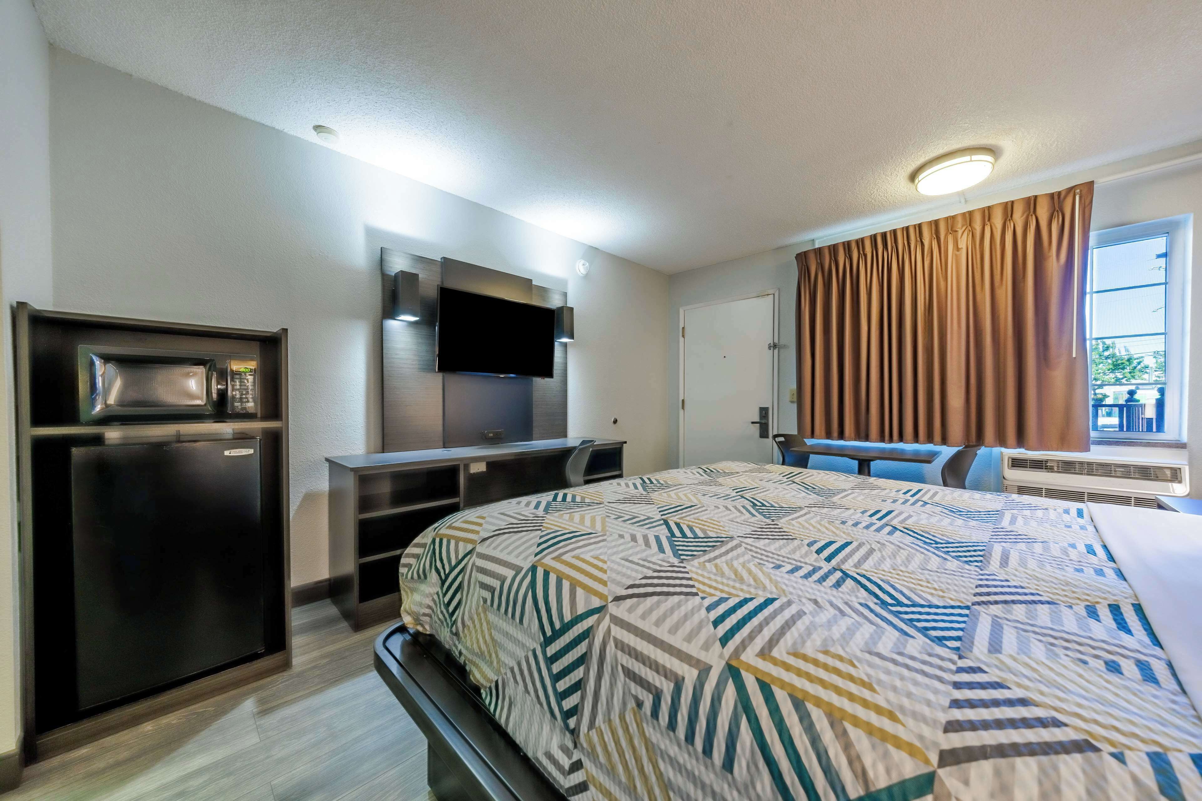 A hotel room with a bed, a TV mounted on the wall, a mini-fridge, a desk with a chair, and a window with curtains.