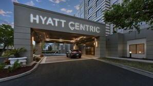 Entrada del hotel Hyatt Centric con un diseño moderno, un coche en la entrada y un cielo despejado al fondo.