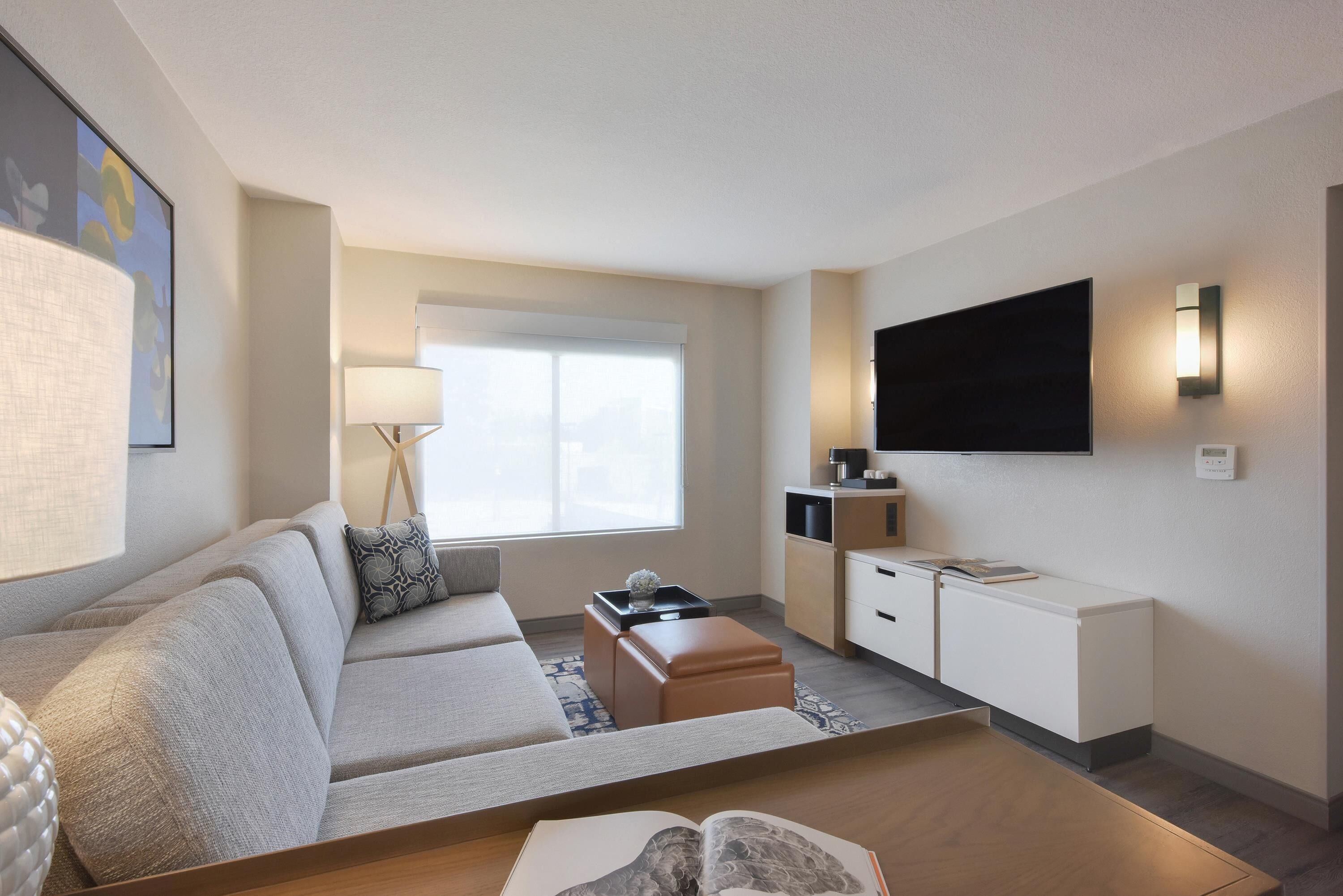 A hotel room with a sofa, a coffee table, a TV, a small kitchenette, and a window with blinds.