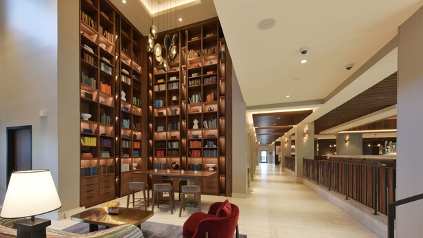 Una biblioteca amplia con altos estantes de madera, una mesa central con sillas y una zona de estar moderna con un sillón rojo.