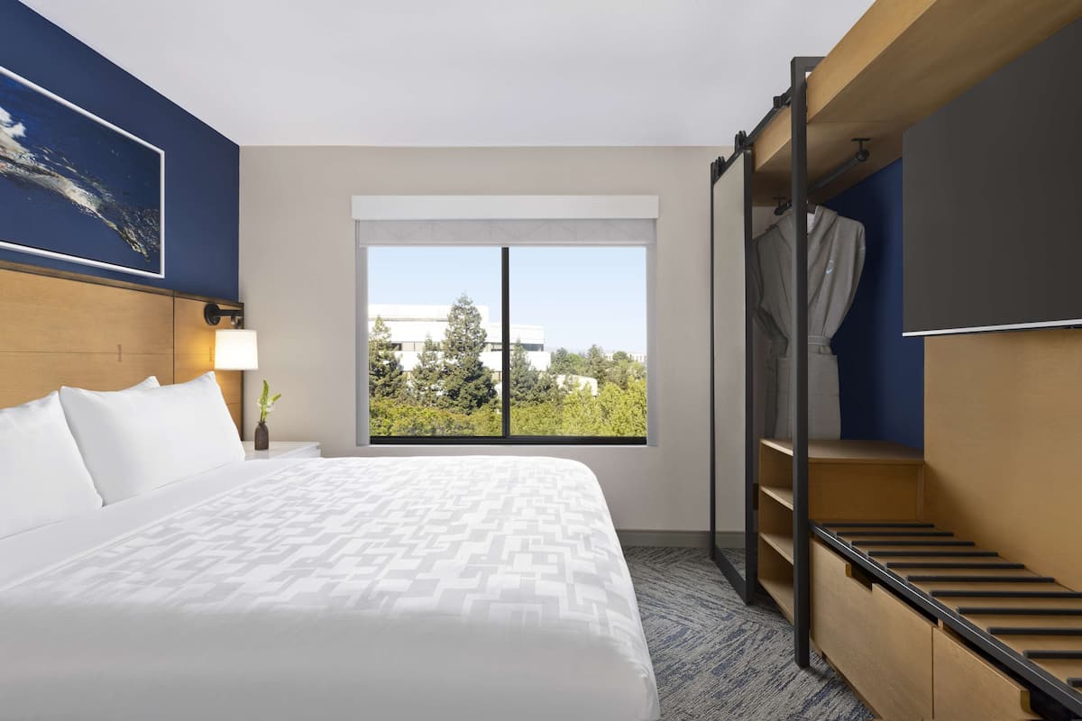 Une chambre d’hôtel avec un grand lit, une fenêtre donnant sur des arbres et des immeubles, un bureau avec une lampe et une armoire intégrée.