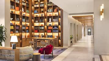 A spacious hallway with a long wooden bookshelf filled with books, a seating area with a red chair, and a table with a lamp.
