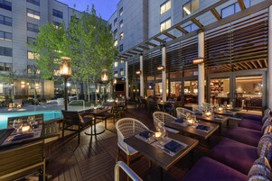 Un área de comedor al aire libre con mesas y sillas, una piscina y un edificio al fondo.