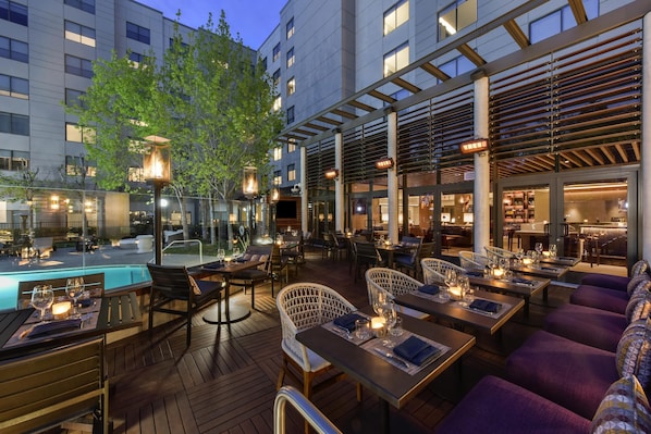 Un área de comedor al aire libre con mesas y sillas, una piscina y un edificio al fondo.