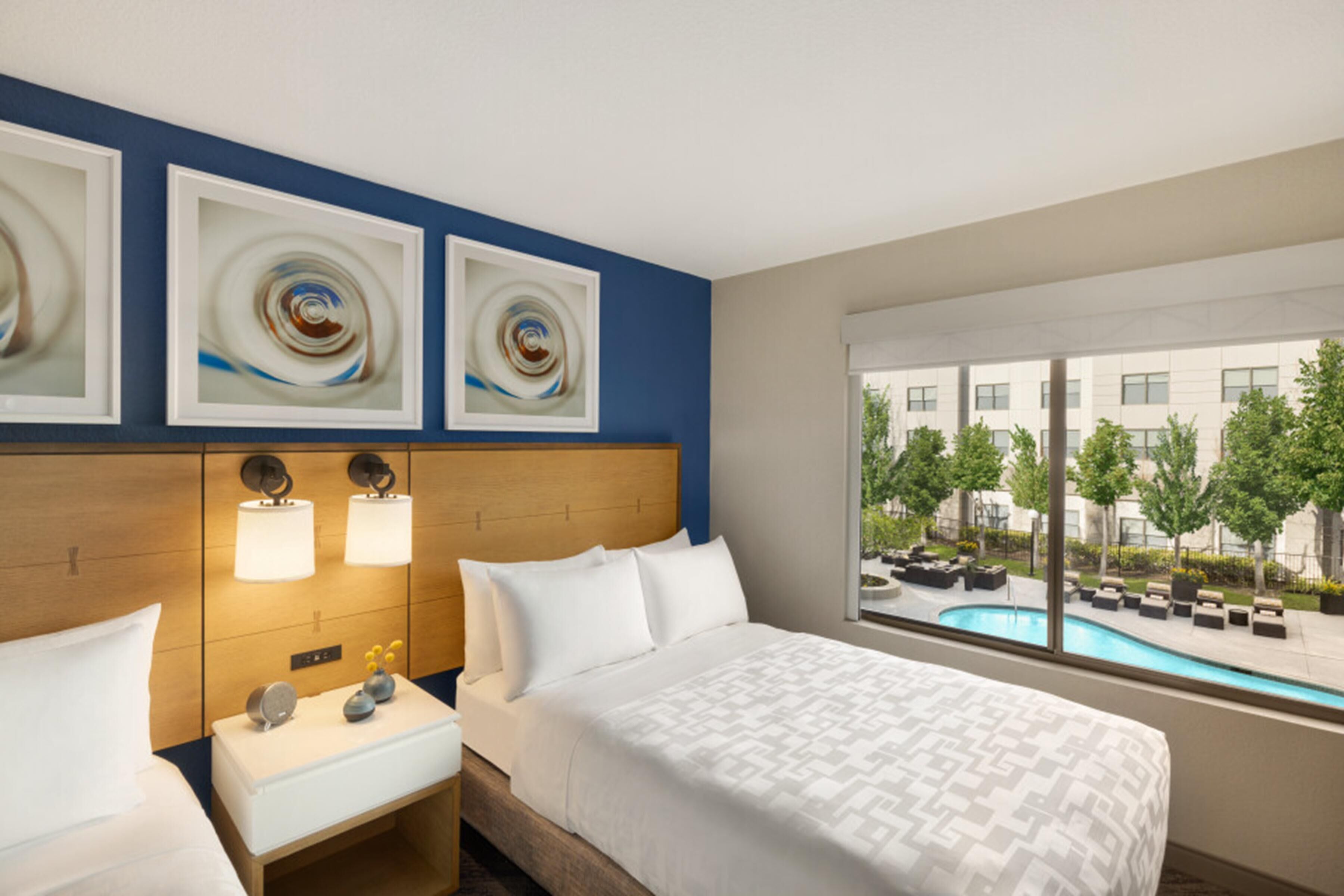 Una habitación de hotel con cama, mesitas de noche, un ventanal con vista a la piscina y una obra de arte abstracta en la pared.