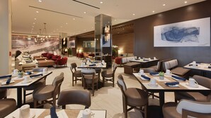 Un moderno vestíbulo de hotel con varias zonas de estar, incluyendo un salón con sofás rojos y un comedor con mesas preparadas para los huéspedes.