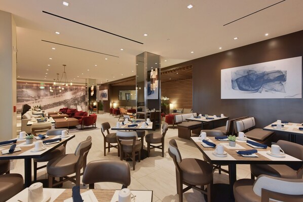 A modern hotel lobby with multiple seating areas, including a lounge with red sofas and a dining area with tables set for guests.
