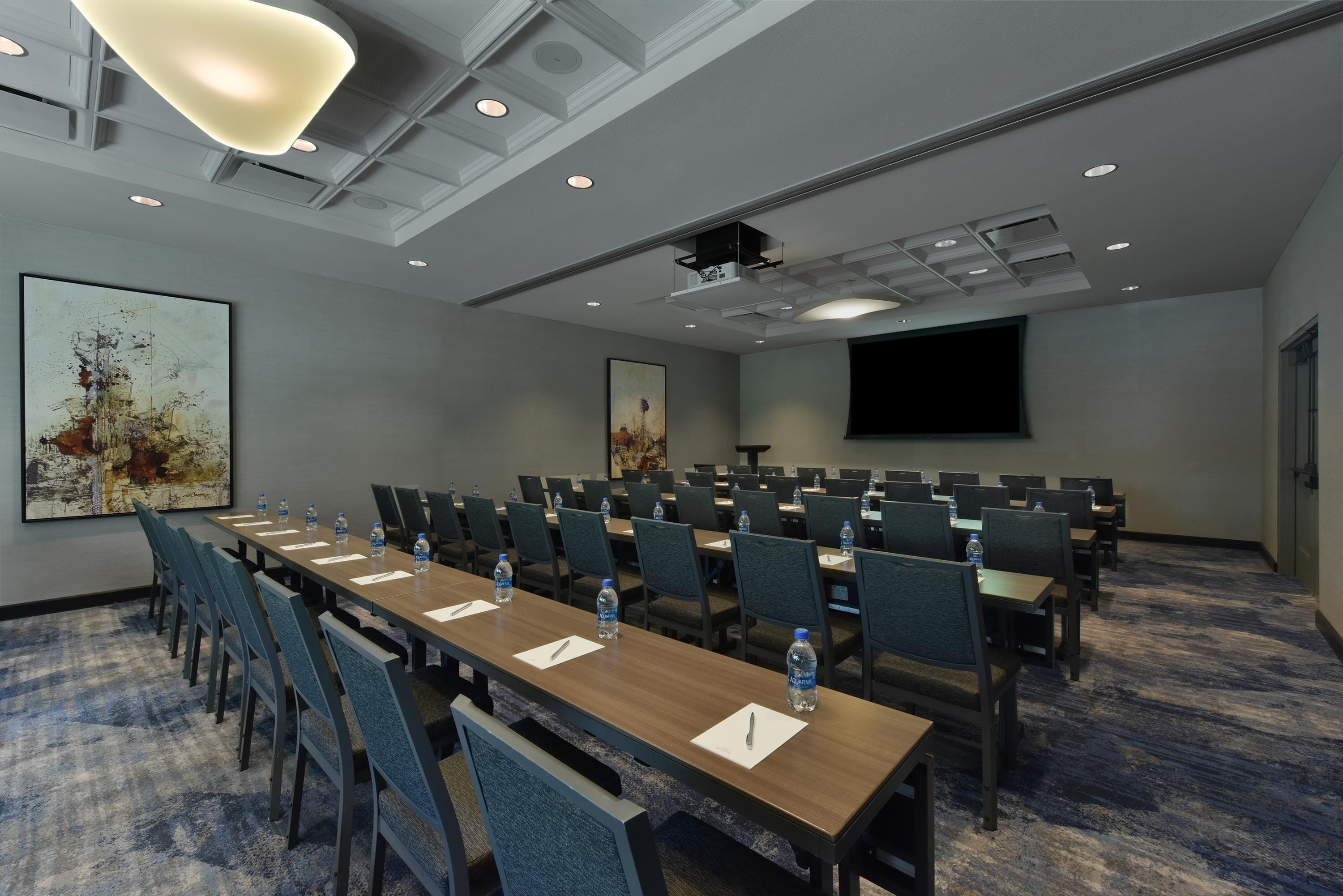 A conference room with rows of tables and chairs, a projector screen, and a large painting on the wall.