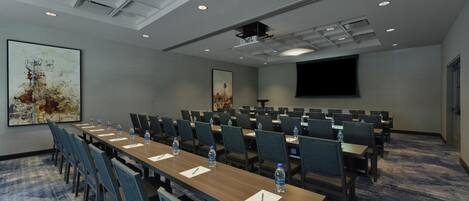 A conference room with rows of tables and chairs, a projector screen, and a large painting on the wall.