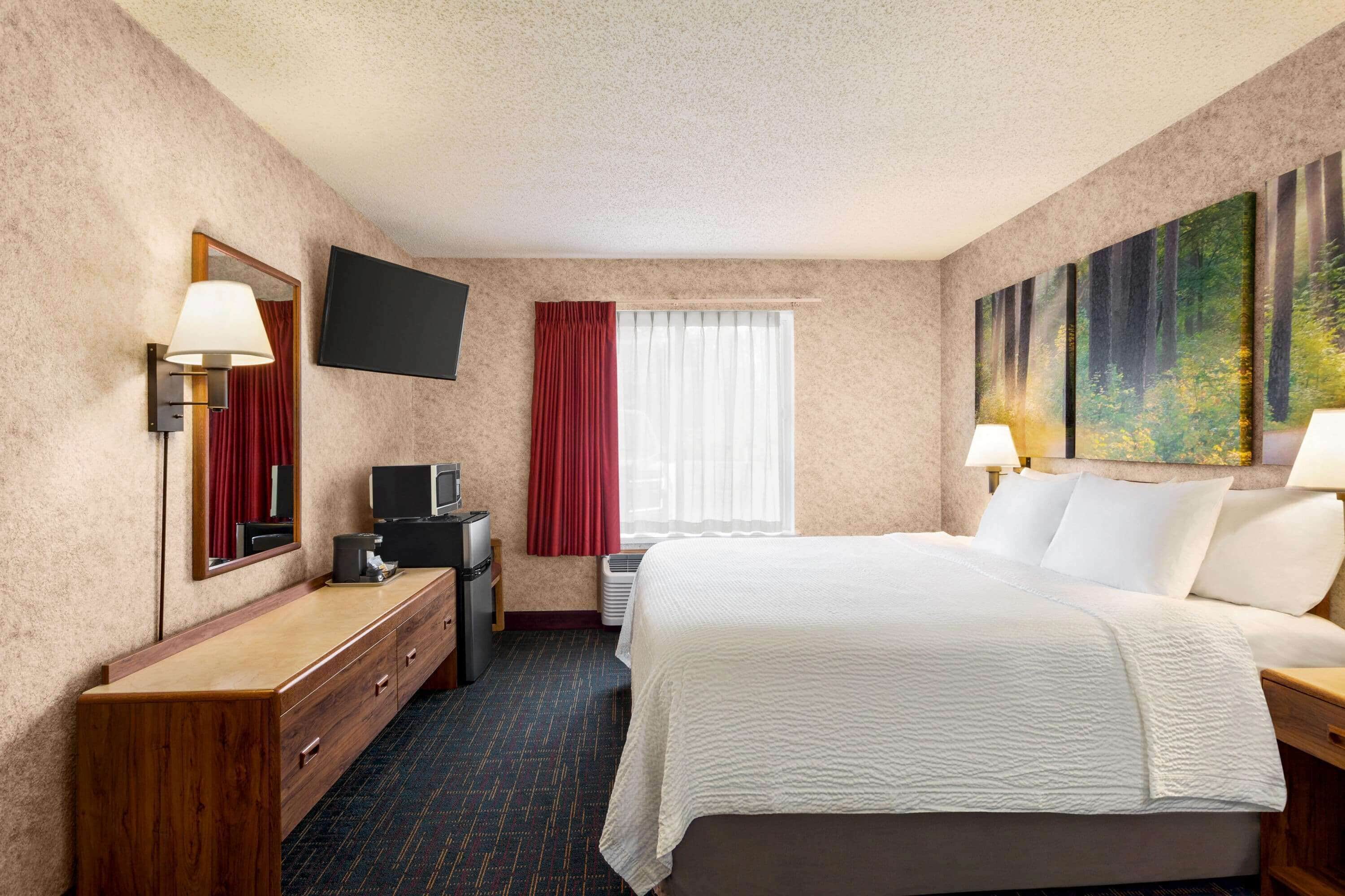 Ein Hotelzimmer mit einem großen Bett, einem Flachbildfernseher, einem Schreibtisch mit Lampe, einem Schminktisch und einem Bild an der Wand.