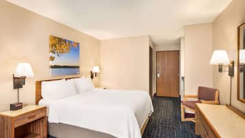 A hotel room with a bed, two bedside lamps, a wooden nightstand, a chair, a desk, and a painting of a lake.