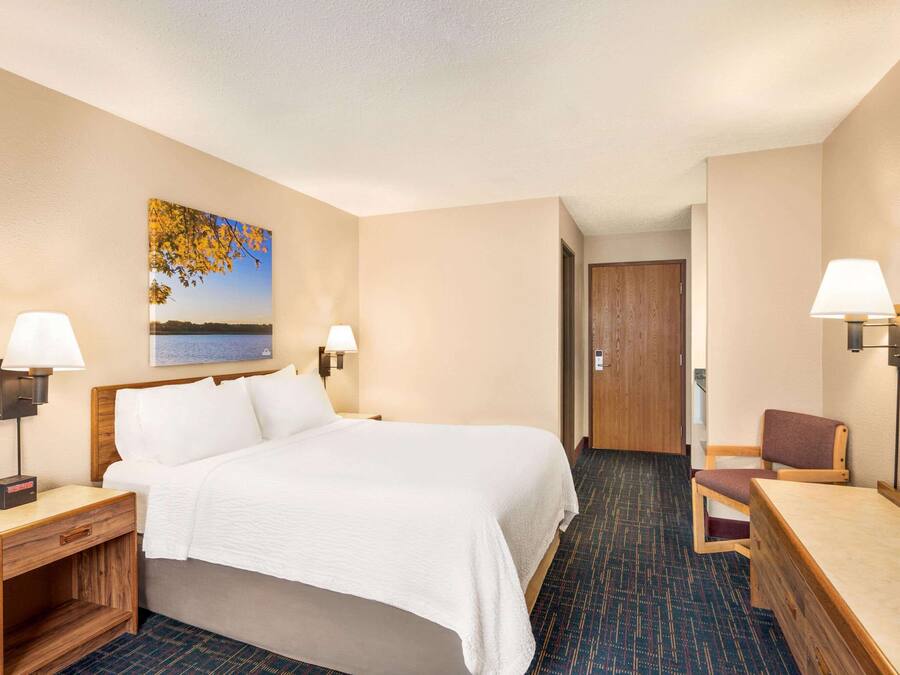 A hotel room with a bed, two bedside lamps, a wooden nightstand, a chair, a desk, and a painting of a lake.