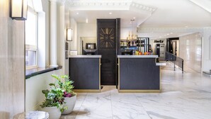 Um hall de hotel com piso de mármore, um balcão de recepção, um relógio e uma escadaria que leva a outro andar.