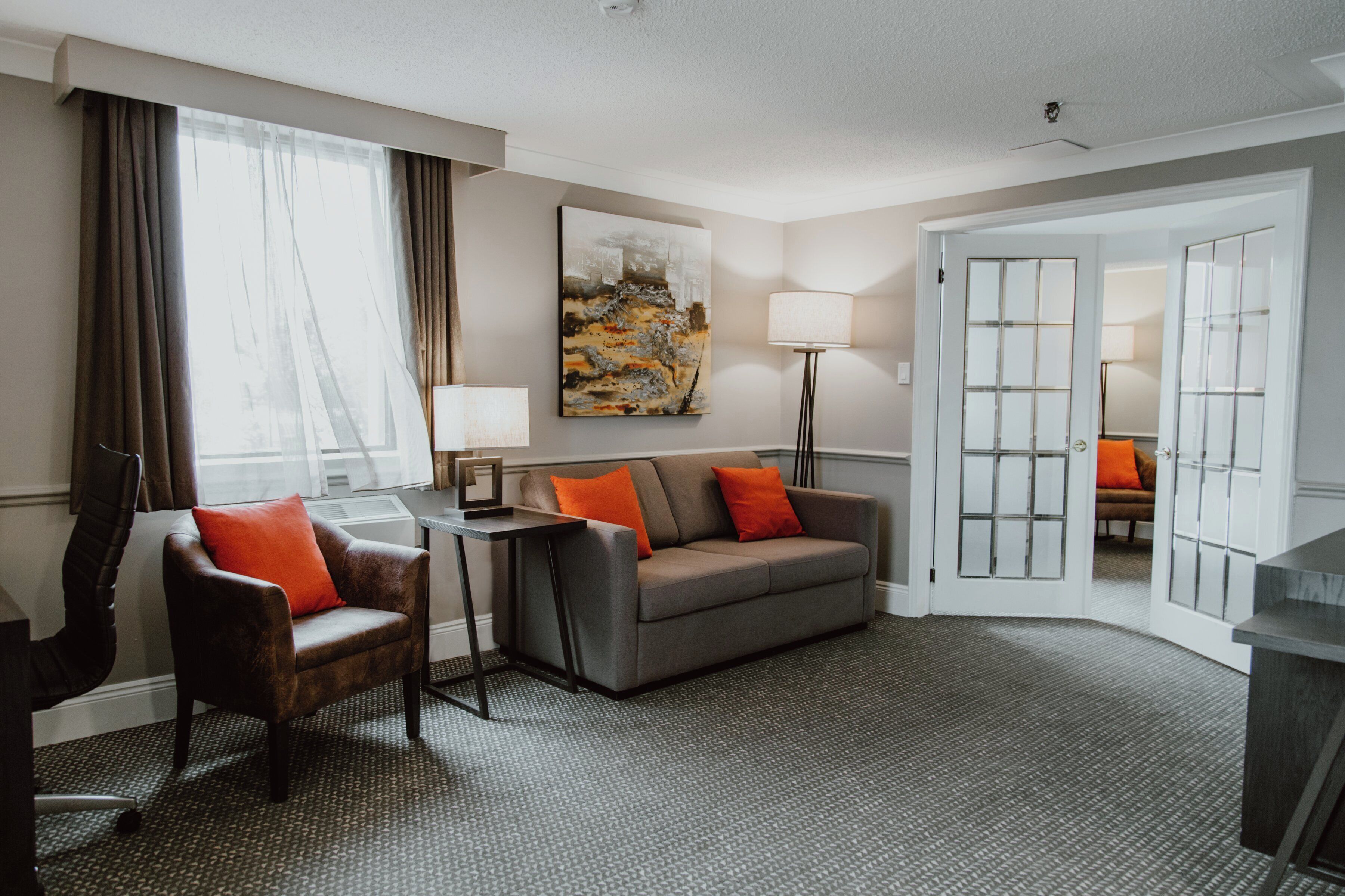 A hotel room with a grey sofa, a brown armchair, a small table, a lamp, and a painting on the wall.