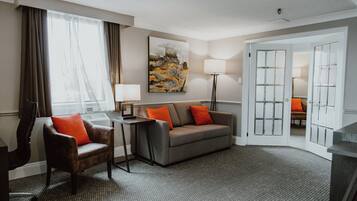A hotel room with a grey sofa, a brown armchair, a small table, a lamp, and a painting on the wall.