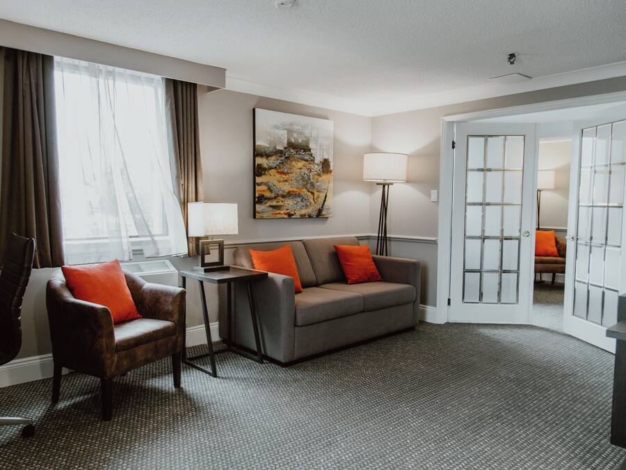A hotel room with a grey sofa, a brown armchair, a small table, a lamp, and a painting on the wall.
